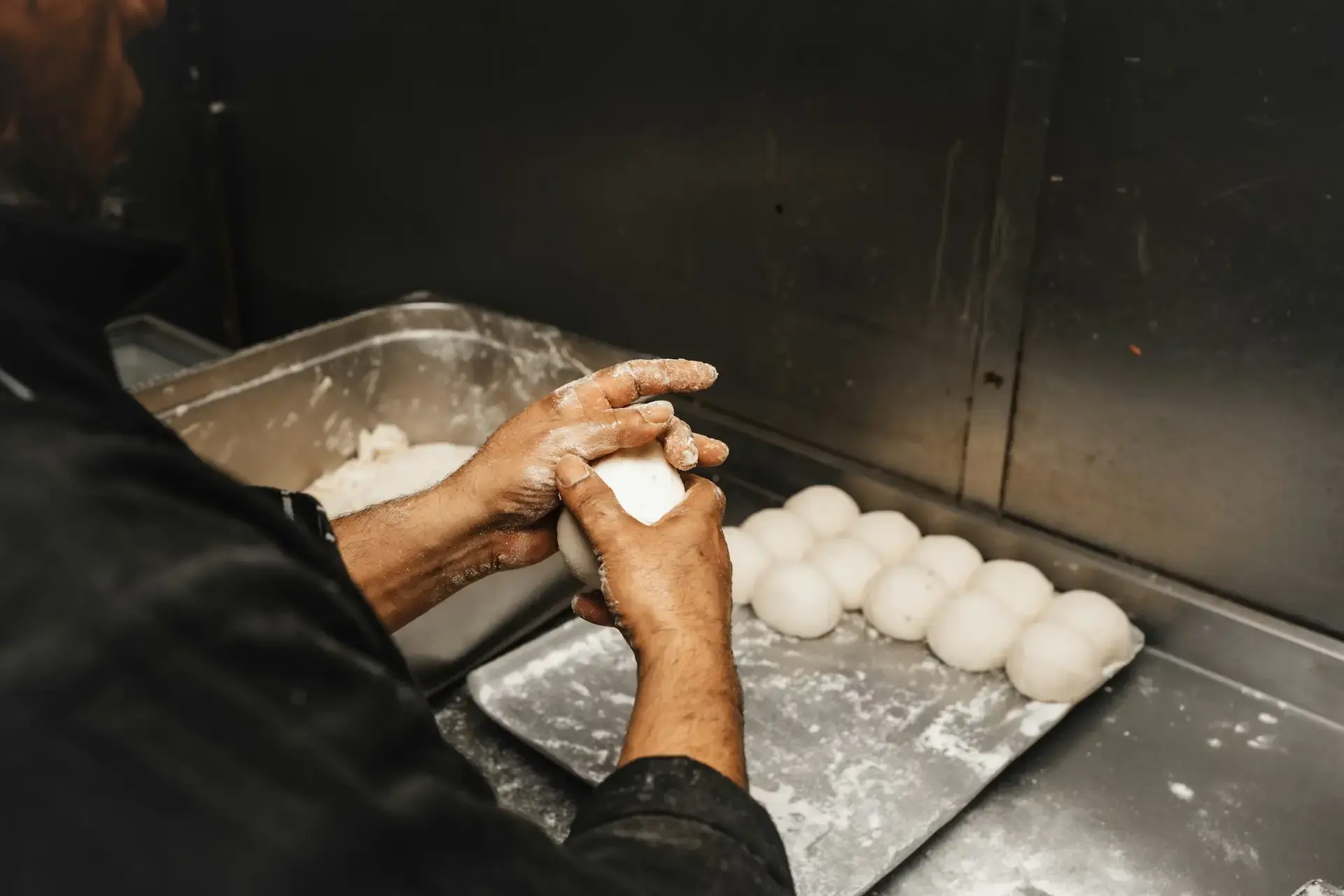 A baker shaping dough by hand in a commercial kitchen, with rows of dough balls resting on a floured tray