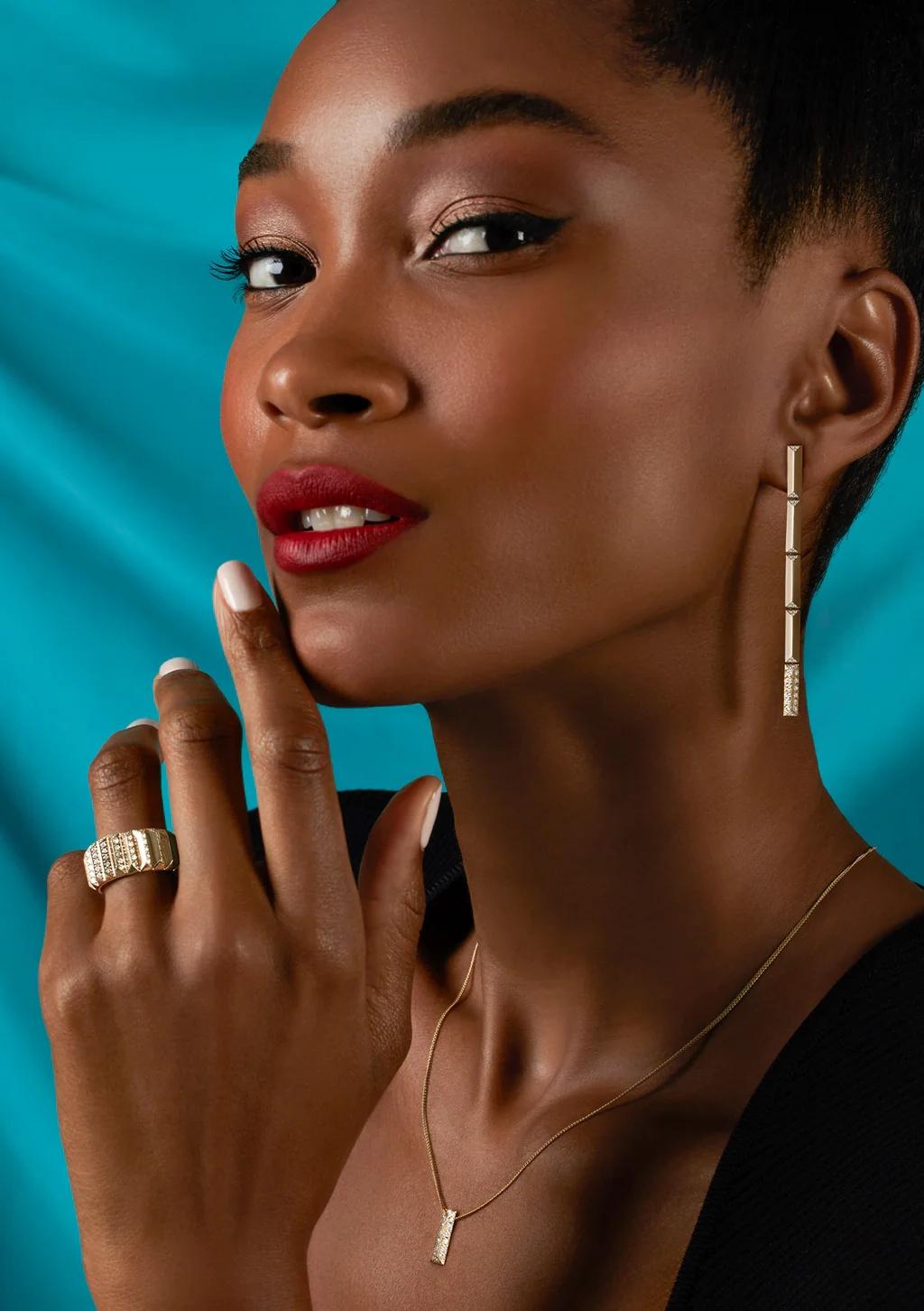 A model wearing CAST gold bar earrings, a pendant necklace and a wide ring against a turquoise backdrop