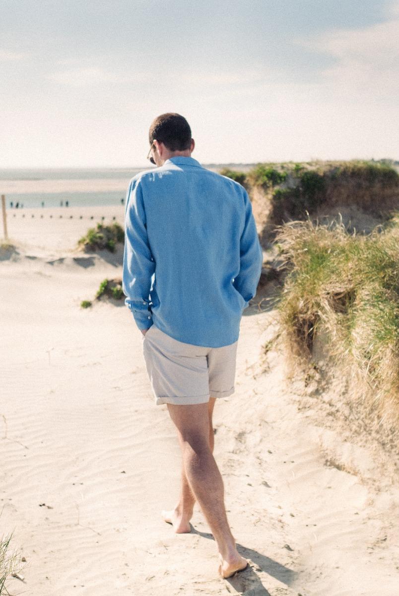 Man in a light blue linen shirt walking barefoot through sand dunes toward the beach