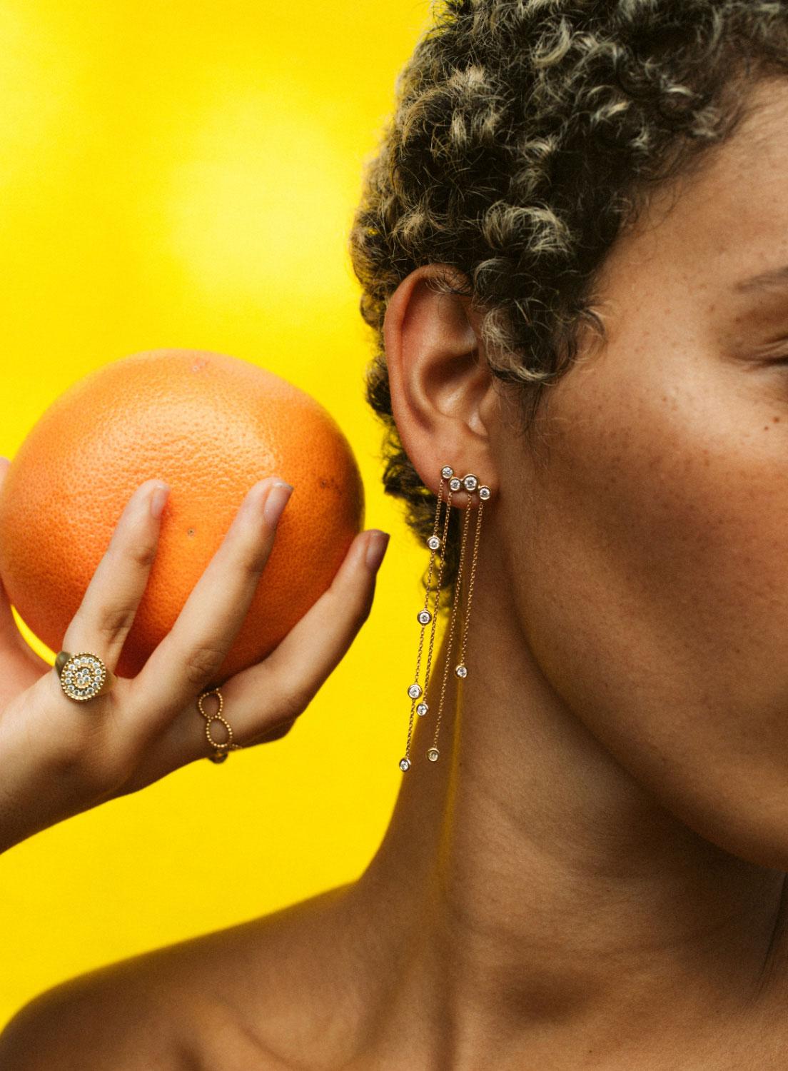 A model wearing Kimai chain drop earrings and diamond rings, holding a grapefruit against a bright yellow background
