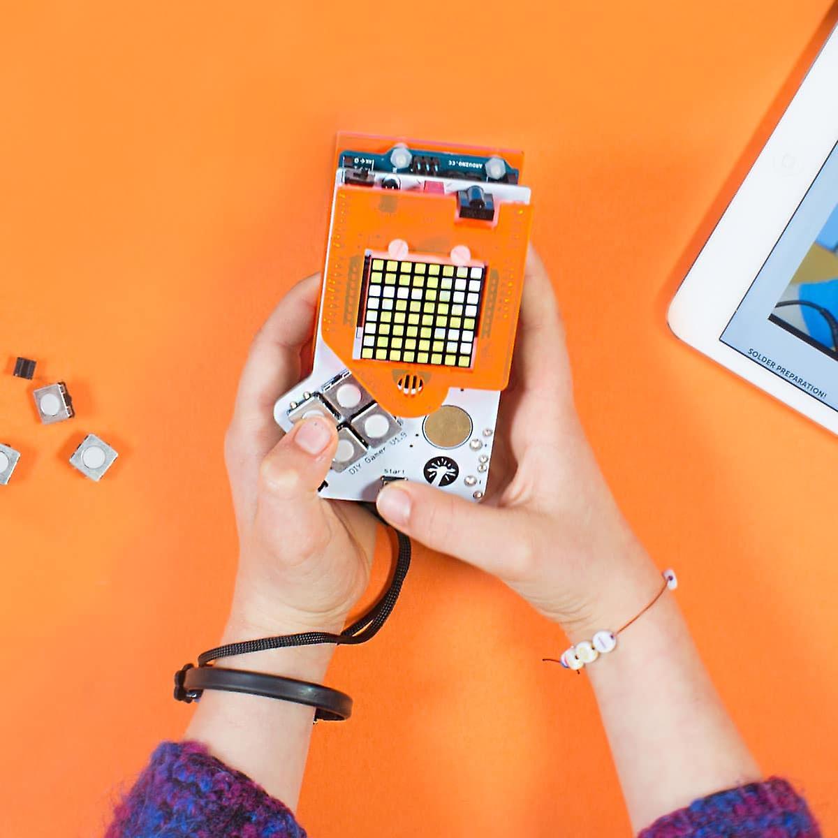 Hands holding a Technology Will Save Us handheld electronics kit with an LED matrix screen, on an orange background