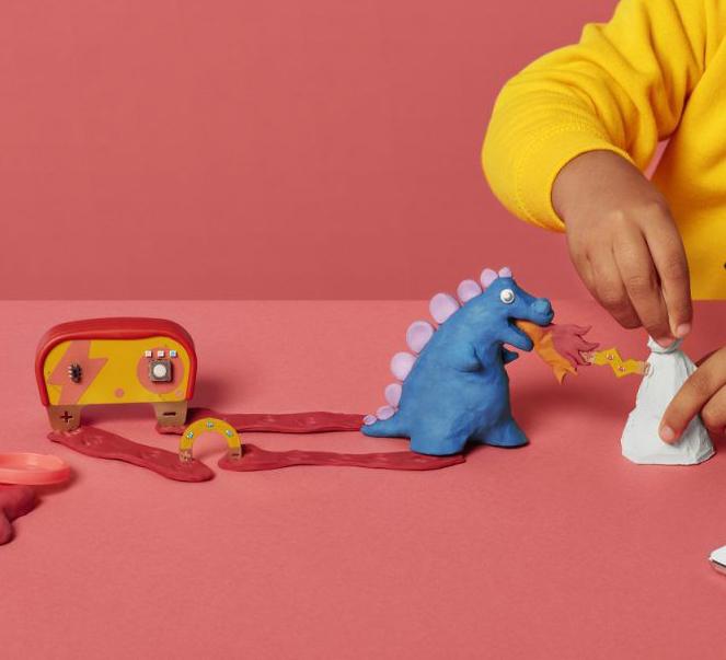Child's hand pressing a clay volcano next to a blue clay dinosaur and a wooden circuit toy, on a pink background