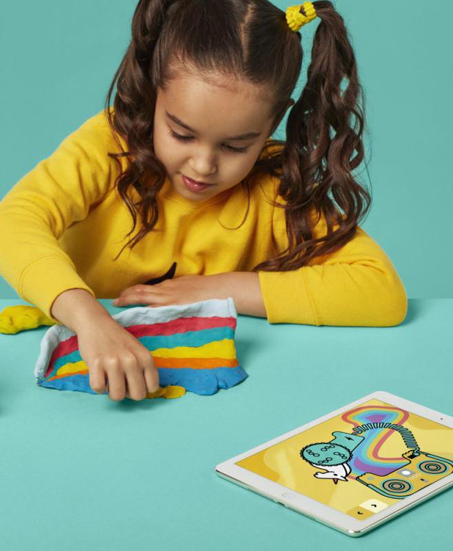 Girl in a yellow sweater shaping colorful modeling clay next to a tablet showing a drawing app